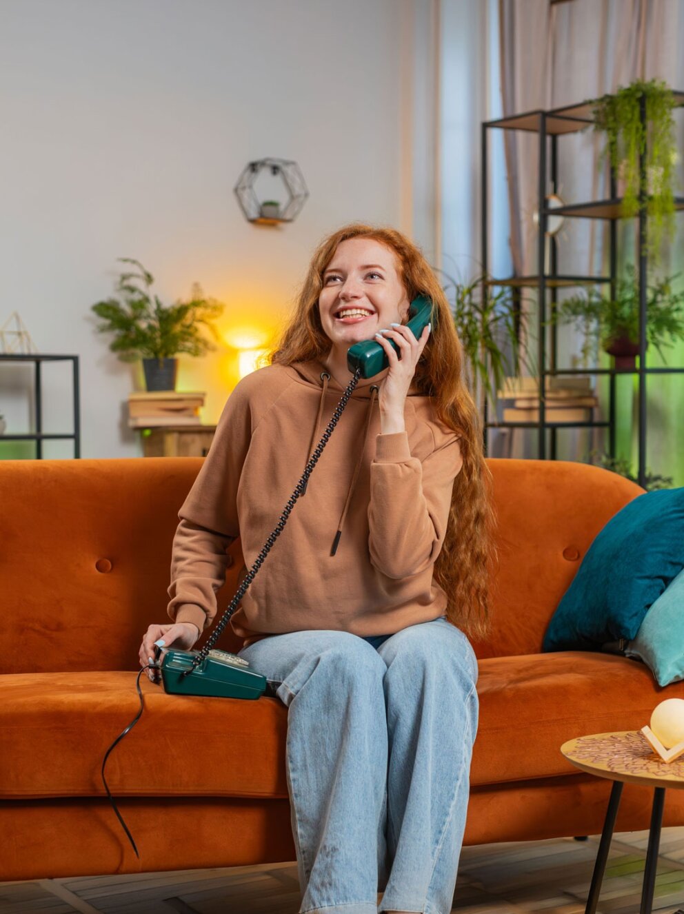 Smiling young woman making wired telephone call conversation with friends sitting on couch at home. Happy excited redhead girl enjoying old-fashioned retro phone from 90s talking in living room.