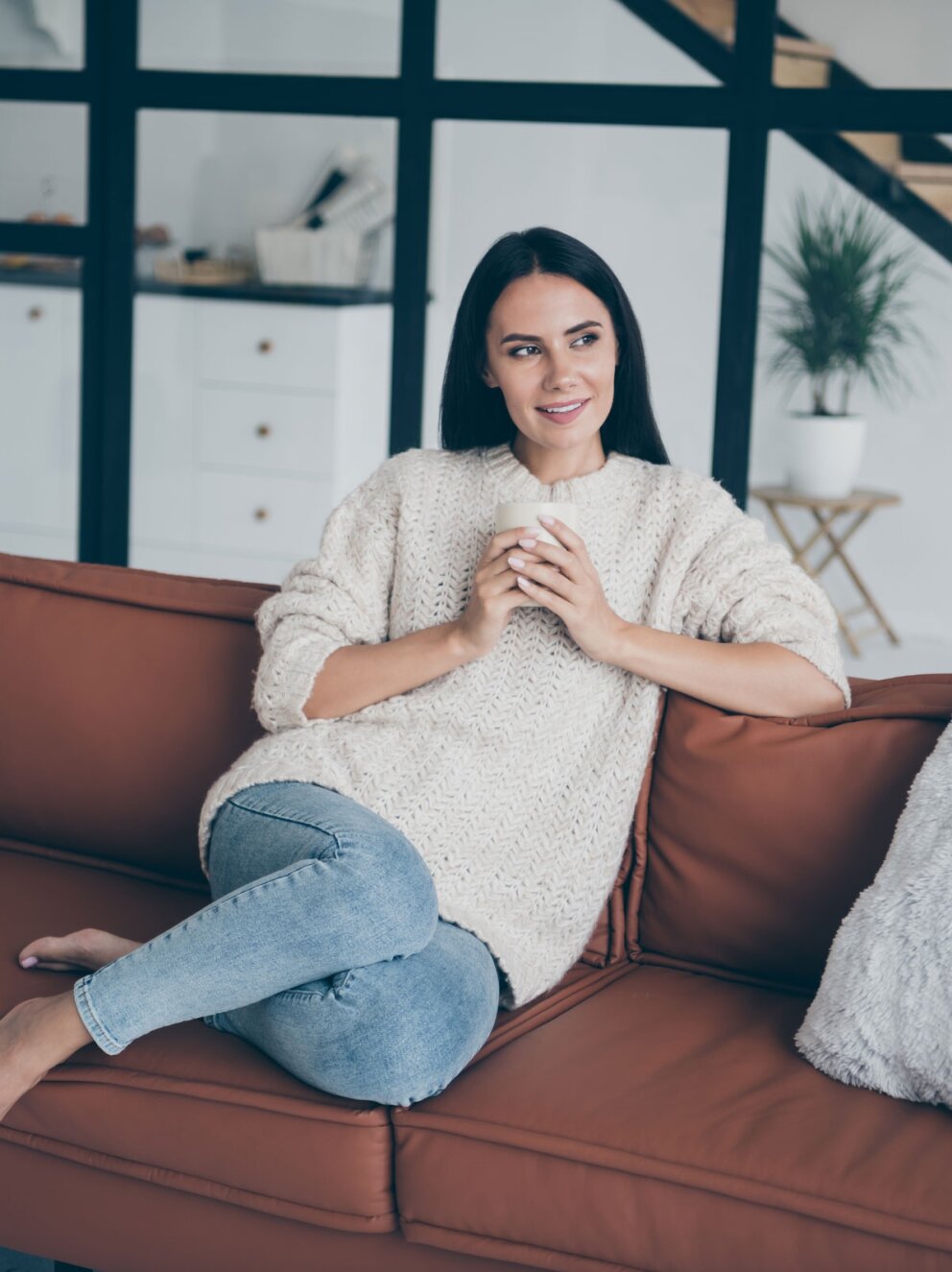 Positive cheerful woman sit on brown leather divan hold cup with latte cappuccino stare window feel inspired imagination dreamy in house indoors